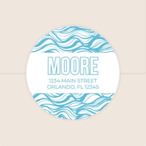 May include: A round white sticker with a blue wave pattern border. The word "MOORE" is in large, light blue block letters. Below, the address "1234 MAIN STREET ORLANDO, FL 12345" is printed in a smaller, light blue font. The sticker is on a light beige background.