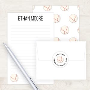 May include: A stationery set featuring a baseball theme. The set includes a lined notepad with the name "ETHAN MOORE" at the top, a patterned sheet with baseballs, an envelope, and a pen. The envelope has a return address label.