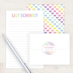 May include: A stationery set featuring a lined notepad, a patterned card, and an envelope. The notepad and card have a pastel heart pattern. The notepad is personalized with the name "LILY SCHMIDT" in rainbow colors. A silver pen rests on the notepad.