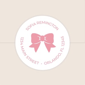 May include: A round, white sticker with a pink bow illustration in the center. The text "SOFIA REMINGTON" arches across the top, with address details and "ORLANDO, FL 12345" around the edge. The sticker is on a light beige background.