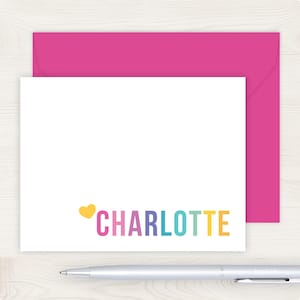 A pink envelope and a white card with the name "Charlotte" written on it, along with a pen, are placed on a wooden surface.