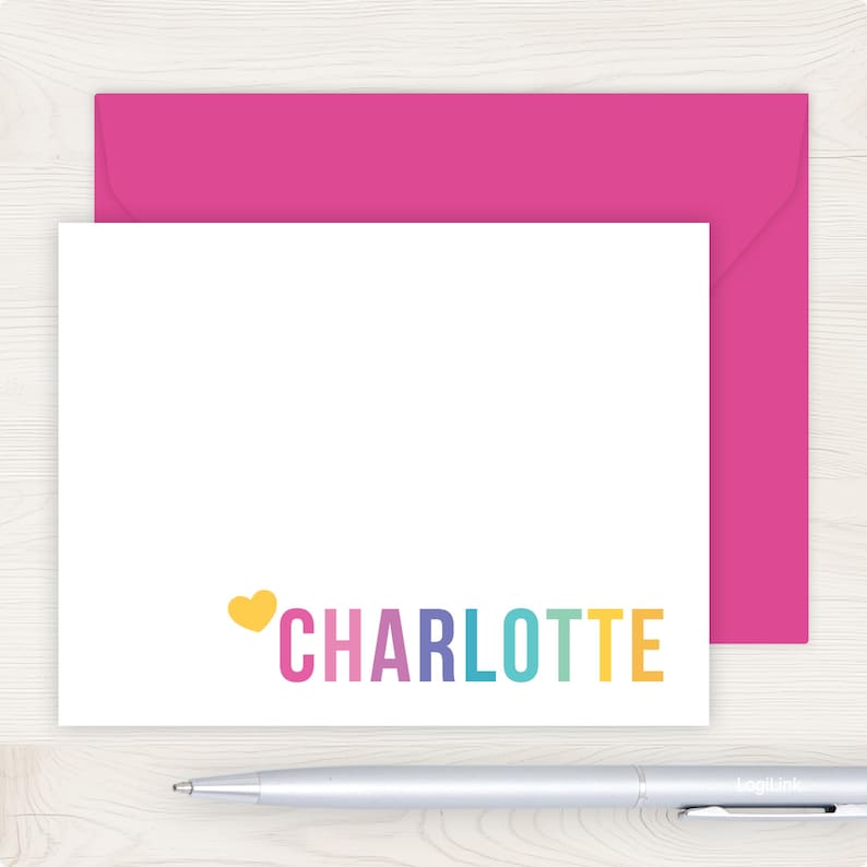 A pink envelope and a white card with the name "Charlotte" written on it, along with a pen, are placed on a wooden surface.