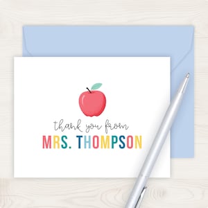 A white card with a colorful graphic of an apple and the text "Thank you from MRS. THOMPSON" written on it, along with a pen on a wooden surface.
