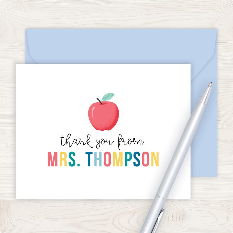 A white card with a colorful graphic of an apple and the text "Thank you from MRS. THOMPSON" written on it, along with a pen on a wooden surface.