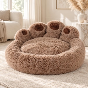 May include: A round, plush pet bed in a light brown color, featuring a high, cushioned border shaped like a large animal paw. The bed includes four distinct, raised toe pads in a darker brown, fuzzy texture, surrounding a soft, circular center.