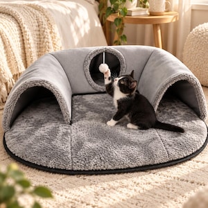 May include: A round, gray pet bed with a plush, textured surface and a curved, tunnel-like structure featuring a circular opening. A small black and white kitten sits on the bed, reaching up to bat at a white pom-pom toy hanging from the top arch.