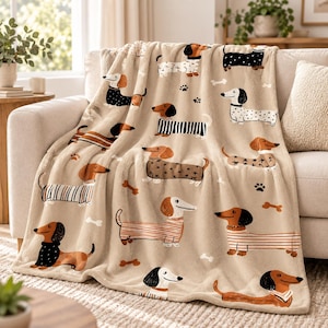 Op de afbeelding: Een zachte beige plaid met een herhalend patroon van teckels in verschillende houdingen en outfits, waaronder gestreepte truien en stippen, afgewisseld met kleine zwarte pootafdrukken en witte botvormen over de gehele stof.