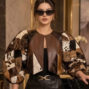 Puede incluir: Una chaqueta corta con un panel central de cuero marrón liso y mangas voluminosas confeccionadas con un mosaico de piel con estampado animal en tonos marrón, negro y blanco. La chaqueta tiene cuello redondo, frente abierto y una silueta estructurada.