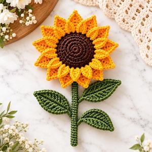 May include: A handcrafted crochet sunflower featuring a dark brown center, vibrant yellow petals, and a green stem with three textured leaves. The flower is constructed with detailed yarn stitching, showcasing a circular head and a long, straight stalk.