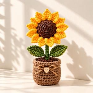 Puede incluir: Un girasol de ganchillo con pétalos de color amarillo brillante y un centro marrón oscuro, sostenido por un tallo verde con dos hojas. La flor está plantada en una maceta de ganchillo de color marrón claro texturizado, decorada con un lazo de cuerda y un pequeño botón de madera en forma de corazón.