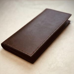 Puede incluir: Una cartera rectangular de cuero marrón oscuro con un acabado liso y mate, y costuras visibles del mismo tono a lo largo de los bordes. La cartera está doblada en un diseño delgado y alargado con un exterior limpio y sin marcas visibles.