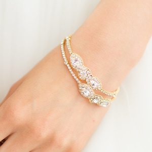 May include: A gold bracelet with clear, sparkling gemstones and a matching ring. The bracelet is worn on a wrist, and the ring is on a finger. The jewelry is set against a white background, suggesting a bridal or formal occasion.