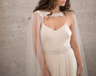 Puede incluir: Un vestido de novia blanco con una capa transparente y un cinturón de cuentas. El vestido tiene un diseño simple y elegante, perfecto para una boda.