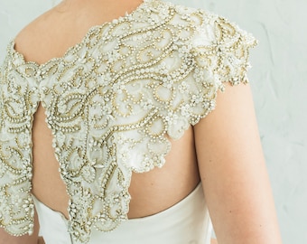 Puede incluir: Un vestido de novia blanco con una superposición de encaje con cuentas en los hombros y la espalda. El vestido tiene una espalda baja y un corpiño ajustado. La novia lleva un peine para el cabello adornado con joyas y pendientes de perlas.