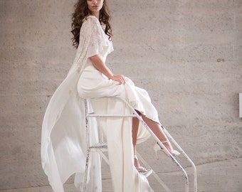 May include: A woman wearing a white gown with a lace overlay sits on a white metal step ladder. The gown has a flowing cape that drapes over the ladder. The woman is looking at the camera.