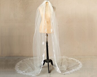 May include: A white bridal veil with a delicate lace trim, displayed on a mannequin against a beige wall.