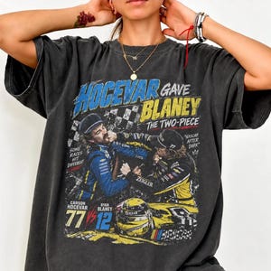 May include: A charcoal gray short-sleeve t-shirt featuring a graphic print of two race car drivers in blue and yellow racing suits. The text reads Hocevar Gave Blaney The Two-Piece, Some Races Hit Different, NASCAR After Dark, Carson Hocevar 77, Ryan Blaney 12.