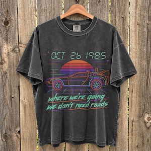May include: A charcoal grey short-sleeve t-shirt featuring a neon-style graphic of a sports car, a sunset, and the text "OCT 26 1985" and "where we're going we don't need roads" printed in a retro digital font style on the front.