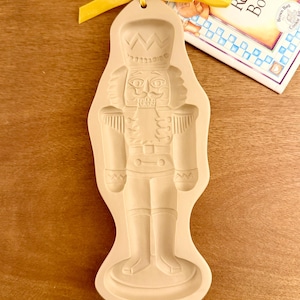 May include: A beige ceramic cookie mold featuring a detailed nutcracker figure with a tall hat, uniform, and boots. The mold has a hole at the top with a bright yellow ribbon tied through it, positioned next to a cookie recipe book.