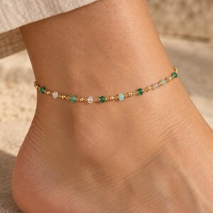 May include: A delicate gold chain anklet featuring a series of small, round, polished gold beads interspersed with tiny, faceted gemstone beads in varying shades of translucent white, light blue, and deep green, creating a subtle, colorful pattern.