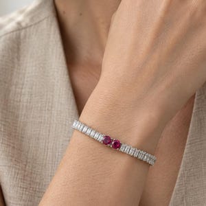 Könnte beinhalten: Ein silberfarbenes Tennisarmband mit einer Reihe rechteckiger, im Baguetteschliff geschliffener, klarer Steine und zwei runden, leuchtend pinkfarbenen Steinen, die nebeneinander in der Mitte angeordnet sind. Das Armband hat ein Metallfinish.