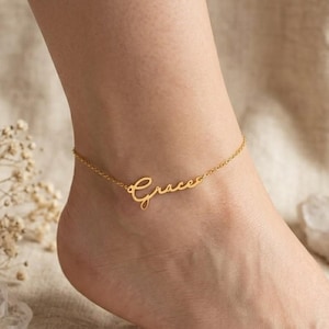 Peut inclure: Une chevillère dorée composée d'une chaîne fine et d'un pendentif central en forme de plaque nominative affichant le prénom Grace dans une écriture cursive. Le bijou est doté d'une chaîne à maillons fins qui s'attache autour de la cheville.