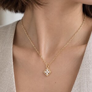 May include: A gold chain necklace featuring a small, diamond-shaped pendant. The pendant has a textured gold border and contains four small, clear, round-cut stones arranged in a flower-like pattern at the center of the diamond shape.