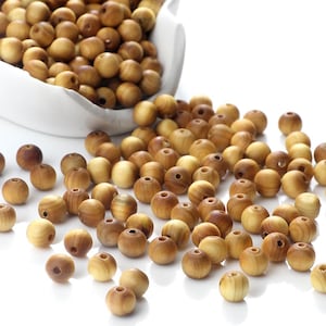 May include: A collection of round, light brown wooden beads with visible natural grain patterns. Many beads are scattered across a white surface, while others are contained within a white ceramic bowl, showing a smooth, polished finish.