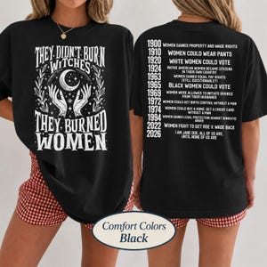 May include: A black t-shirt featuring white graphic text. The front displays hands holding a crescent moon with the text "THEY DIDN'T BURN WITCHES THEY BURNED WOMEN." The back lists historical milestones for women from 1900 to 2026.