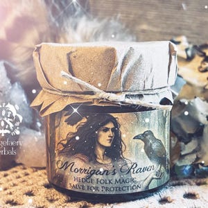 May include: A small glass jar of Hedge Folk Magic salve for protection. The label features an illustration of a woman and a raven, with the text "Morrigan's Raven". The jar is sealed with brown paper and twine.