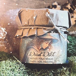 May include: A small, round jar of salve with a paper lid tied with twine. The jar features a label with the words "Wood Witch" and an illustration of a woman in a witch hat. The text "Hedge Folk Magic Salve for Healing" is also on the label.