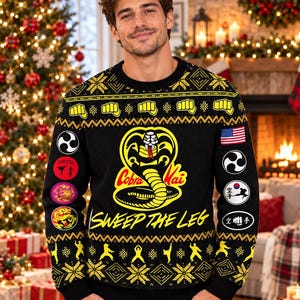 Puede incluir: Un suéter de punto negro de manga larga con el logotipo amarillo de Cobra Kai y el texto SWEEP THE LEG. El diseño incluye iconos de puños amarillos, patrones geométricos y parches circulares en las mangas con símbolos de artes marciales.