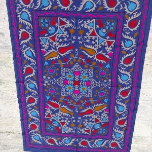 May include: A rectangular blue textile featuring intricate hand-stitched embroidery. The design includes a central star motif surrounded by floral and leaf patterns in vibrant shades of red, pink, light blue, and gold, framed by a border.