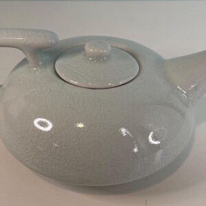 May include: A light blue ceramic teapot with a rounded body, a curved handle, and a spout. The teapot has a lid with a small knob and a textured surface. The teapot is designed for brewing tea.
