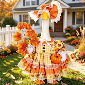 May include: A white goose figurine wearing an orange and yellow gingham dress with lace trim, a matching bonnet, and a white ribbon bow. The goose holds a cluster of orange maple leaves in one wing and a small orange pumpkin in the other wing.