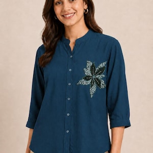 Può includere: Una camicia abbottonata color verde ottanio scuro con colletto alla coreana, maniche a tre quarti e un'applicazione floreale sul petto sinistro. L'applicazione è composta da petali in tessuto verde scuro e ottanio con perline dettagliate.