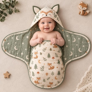 May include: A baby swaddle blanket featuring a hood with fox ears and a face design. The fabric is cream-colored with a pattern of foxes, trees, mushrooms, and moons. The interior lining is sage green with a white moon and star print pattern.
