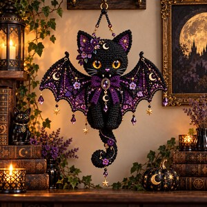 Puede incluir: Un gato negro tejido a ganchillo con grandes ojos naranjas, colmillos y amplias alas de murciélago. La figura está adornada con flores moradas, dijes de media luna dorados, estrellas colgantes y cristales morados facetados.