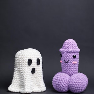 May include: Two crocheted plush figures. One is a white ghost with black yarn eyes and a mouth. The other is a light purple phallic-shaped figure with a rounded top, two rounded base sections, black bead eyes, a stitched smile, and pink cheeks.