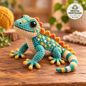May include: A crochet lizard toy featuring a teal body with yellow spots, a cream-colored underbelly, and an orange spiky crest along its back. The lizard has large black eyes and a striped tail. The image includes text: DIGITAL DOWNLOAD NOT A PHYSICAL ITEM.