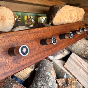 May include: A rectangular wooden wall rack featuring four hooks made from black and silver shotgun shell casings. The wood has a smooth, polished brown finish, and the metal shell casings are mounted horizontally along the front surface.