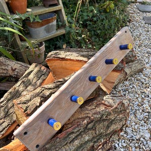 May include: A rectangular wooden wall rack featuring five hooks made from blue and gold shotgun shell casings. The light brown wood has a visible grain pattern, and the rack includes pre-drilled mounting holes at both ends for secure installation.