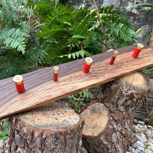 May include: A rectangular wooden wall rack featuring five red shotgun shell casings used as hooks. The board displays a natural wood grain with a dark brown finish on one side and a lighter brown tone on the other, with a small hole at each end.