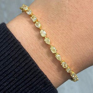 Peut inclure: Un bracelet tennis en or composé d'une rangée continue de pierres jaune clair en forme de poire serties dans une monture métallique. Le bracelet est porté au poignet, mettant en valeur l'agencement uniforme des gemmes facettées et la monture en or.