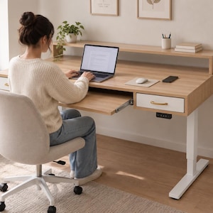 Pode incluir: Uma secretária ajustável em altura de madeira clara com estrutura branca, apresentando uma prateleira elevada para monitor, uma bandeja deslizante para teclado e duas gavetas brancas com puxadores dourados e um painel de controlo digital.
