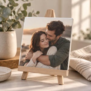 May include: A square ceramic tile featuring a printed photograph of two people embracing outdoors at sunset, displayed on a small wooden easel. The image shows a person with long dark hair wearing a cream sweater hugging another person.