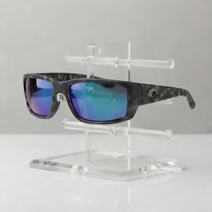 May include: Sunglasses with a dark grey and black mottled frame and reflective blue-tinted lenses, resting on a clear acrylic multi-tier display stand. The frame features a small silver logo detail on the temple and clear nose pads.