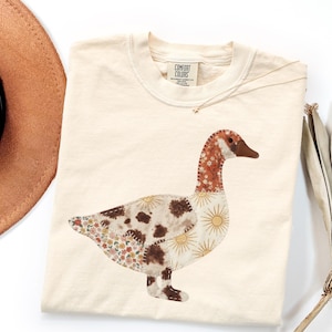 以下が含まれることがあります： 前面に大きなガチョウのグラフィックが描かれたクリーム色のTシャツ。ガチョウは花柄、太陽の光、茶色の牛柄の様々な布パッチで構成されており、すべて目に見えるステッチで縁取られています。首元のラベルにはComfort Colorsと記載されています。