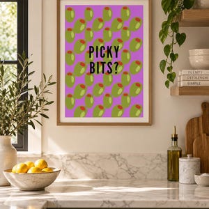 May include: A rectangular wall art print in a light wood frame featuring a repeating pattern of green olives with red pimentos on a bright purple background. The text PICKY BITS? is printed in bold black sans-serif letters in the center.