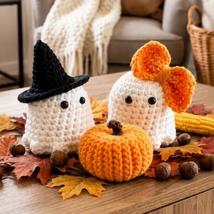 May include: Two white crocheted ghost figures and one orange crocheted pumpkin. One ghost wears a black pointed hat, and the other ghost features a large orange bow. The figures are arranged with scattered brown autumn leaves and an acorn.